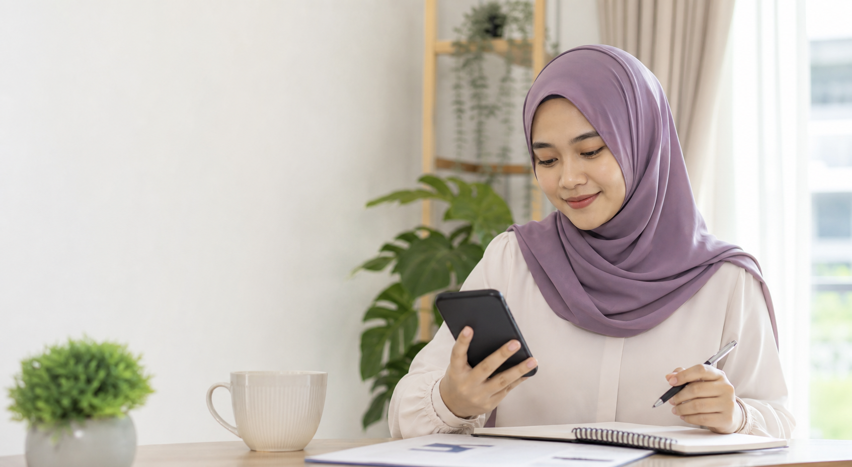 Seorang wanita Malaysia menyemak bajet peribadi menggunakan telefon dan buku nota